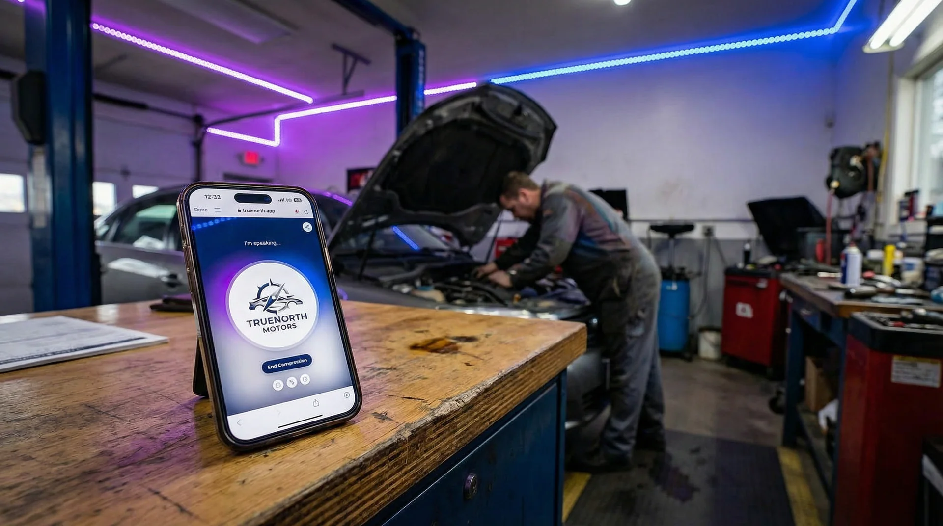 Auto mechanic working under a car hood while a phone sits unanswered on a workbench
